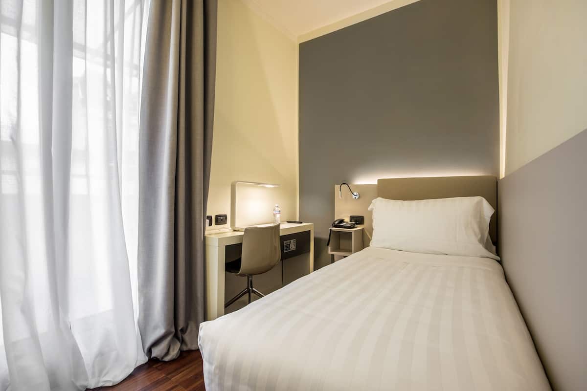 A hotel room with a bed, desk, chair, and a nightstand with a lamp.