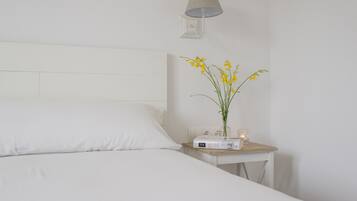 A neatly made bed with white linens, a bedside table with a lamp, a book, and a vase with yellow flowers.