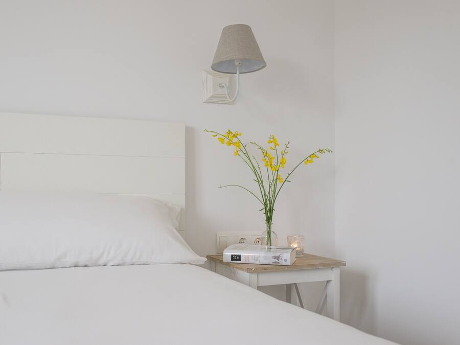 A neatly made bed with white linens, a bedside table with a lamp, a book, and a vase with yellow flowers.