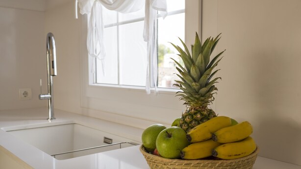Una cocina con un lavamanos blanco, una ventana con cortinas blancas y una cesta de frutas que incluye manzanas, plátanos y una piña.