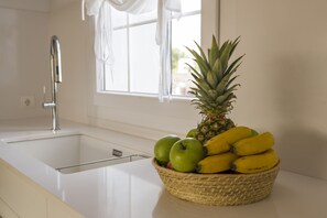 Una cocina con un lavamanos blanco, una ventana con cortinas blancas y una cesta de frutas que incluye manzanas, plátanos y una piña.