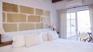 Habitación de hotel con una cama grande, pared de piedra, balcón con una silla y unidad de aire acondicionado.