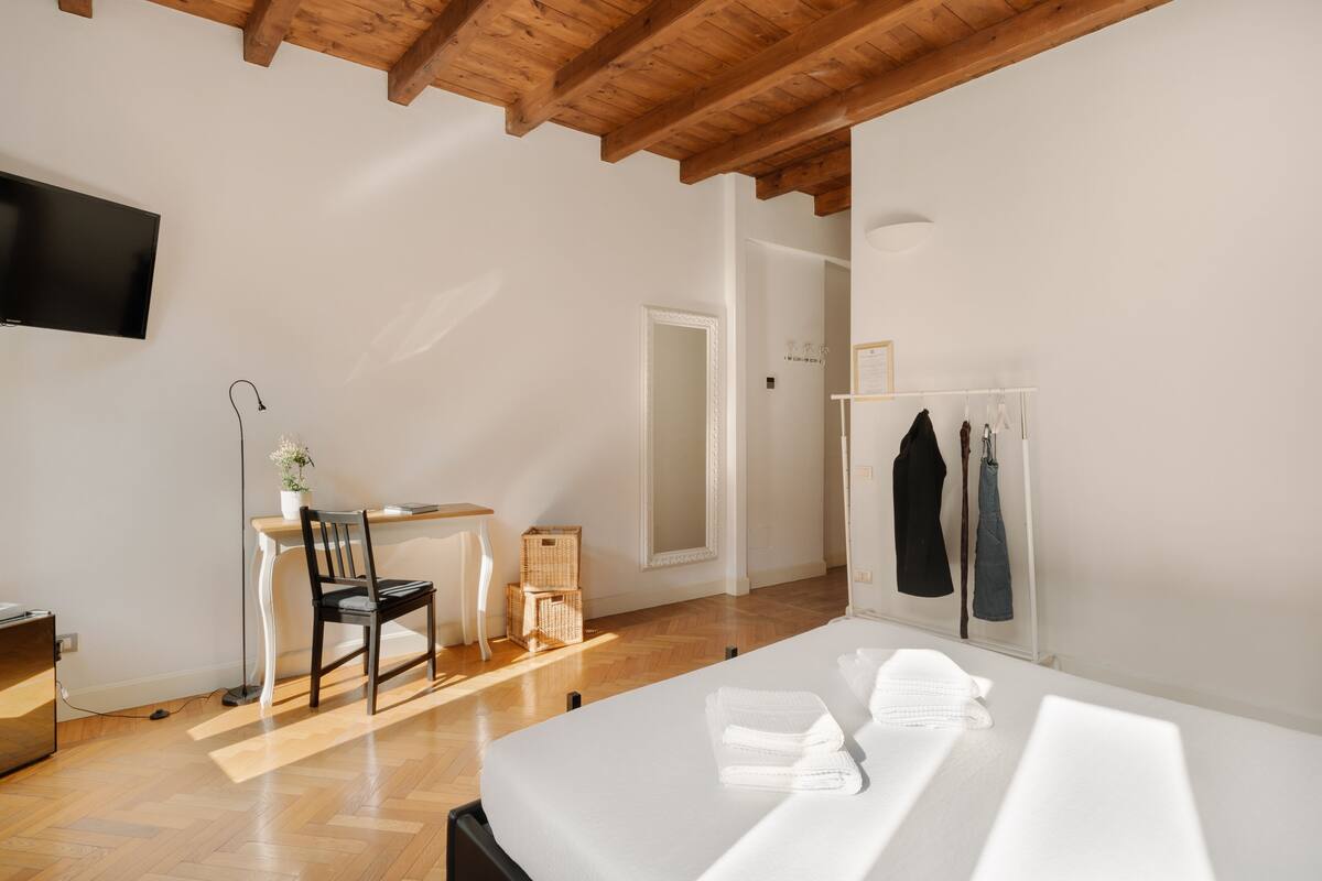 Habitació d'hotel moderna amb sostre de fusta, televisió de pantalla plana penjada a la paret, escriptori amb cadira, llit amb tovalloles ordenades i armari amb roba penjada.
