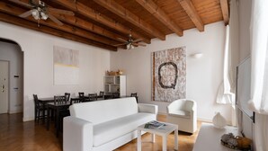 A modern living room with a white sofa, dining table, and chairs. There is a ceiling fan, wooden beams, and artwork on the walls. - Quarenghi16 (Bergamo)