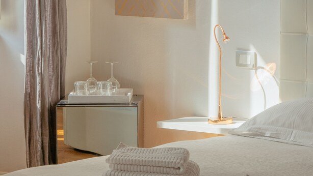 A neatly made bed with folded towels, a bedside table with a lamp, and a wall-mounted light fixture. - Quarenghi16 (Bergamo)
