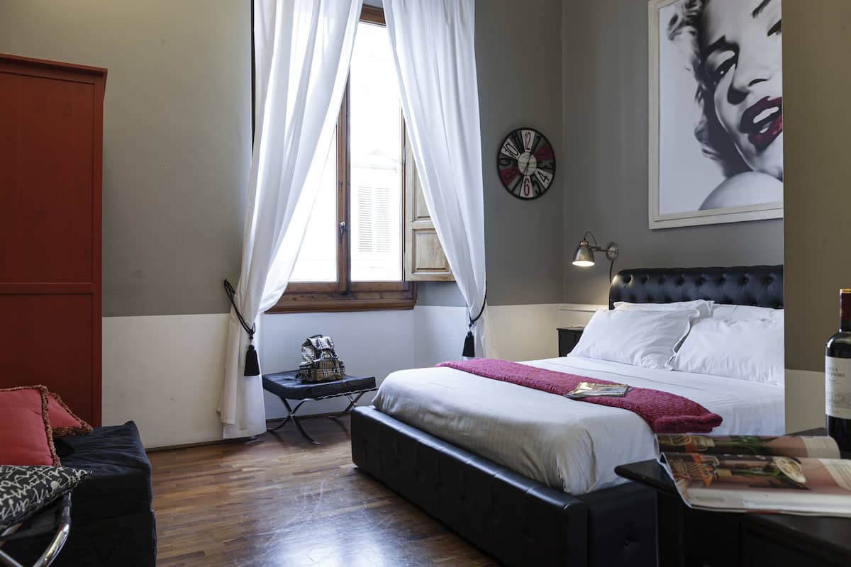 Une chambre à coucher avec un lit, une table de chevet, une lampe, une horloge et une fenêtre avec des rideaux.