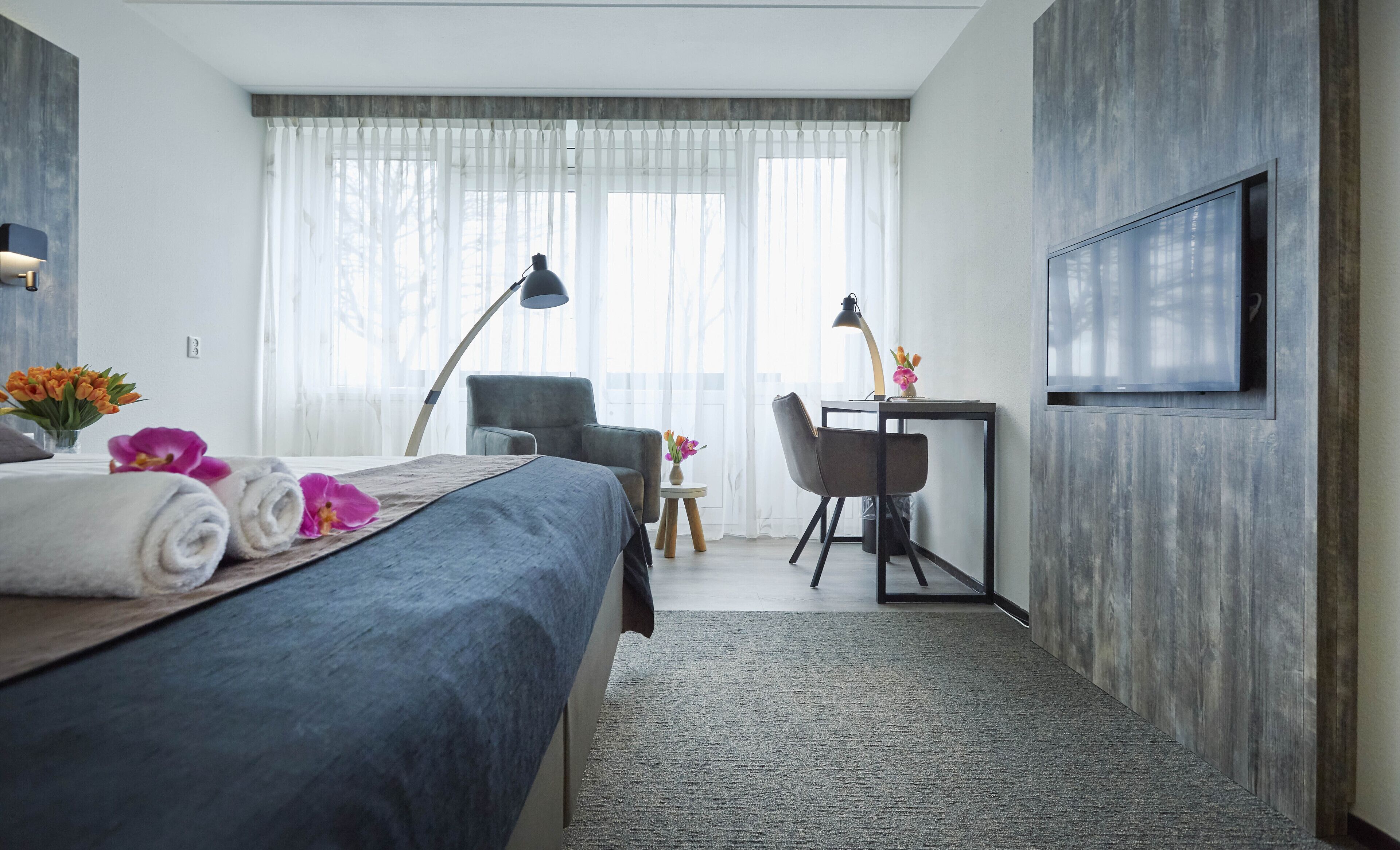 Habitación de hotel con cama, escritorio, silla, televisor y ventana con cortinas translúcidas.