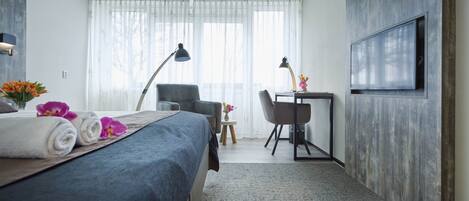 Habitación de hotel con cama, escritorio, silla, televisor y ventana con cortinas translúcidas.