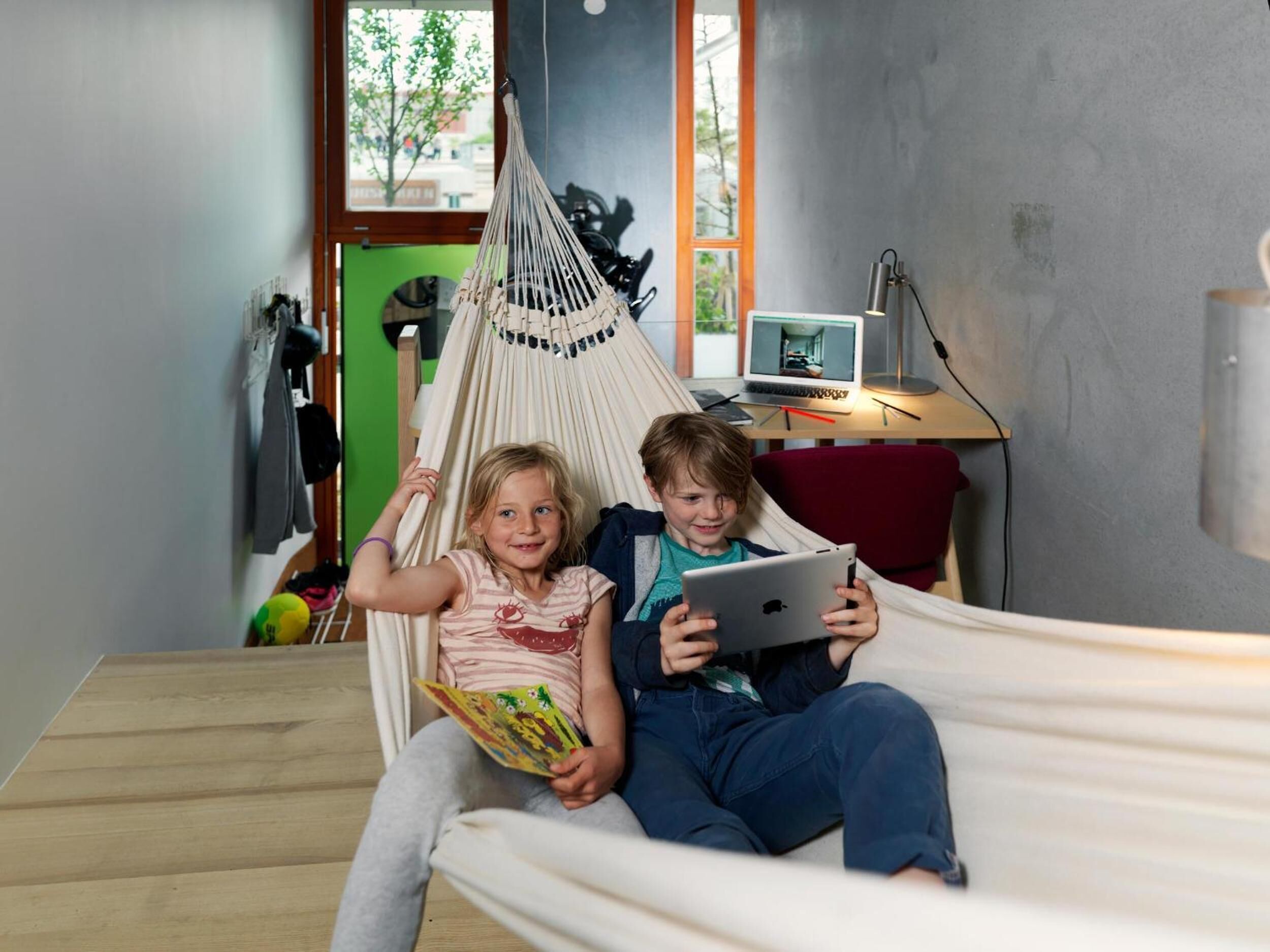 Two children in a hammock in a room with a desk, laptop, and a view of the outdoors.