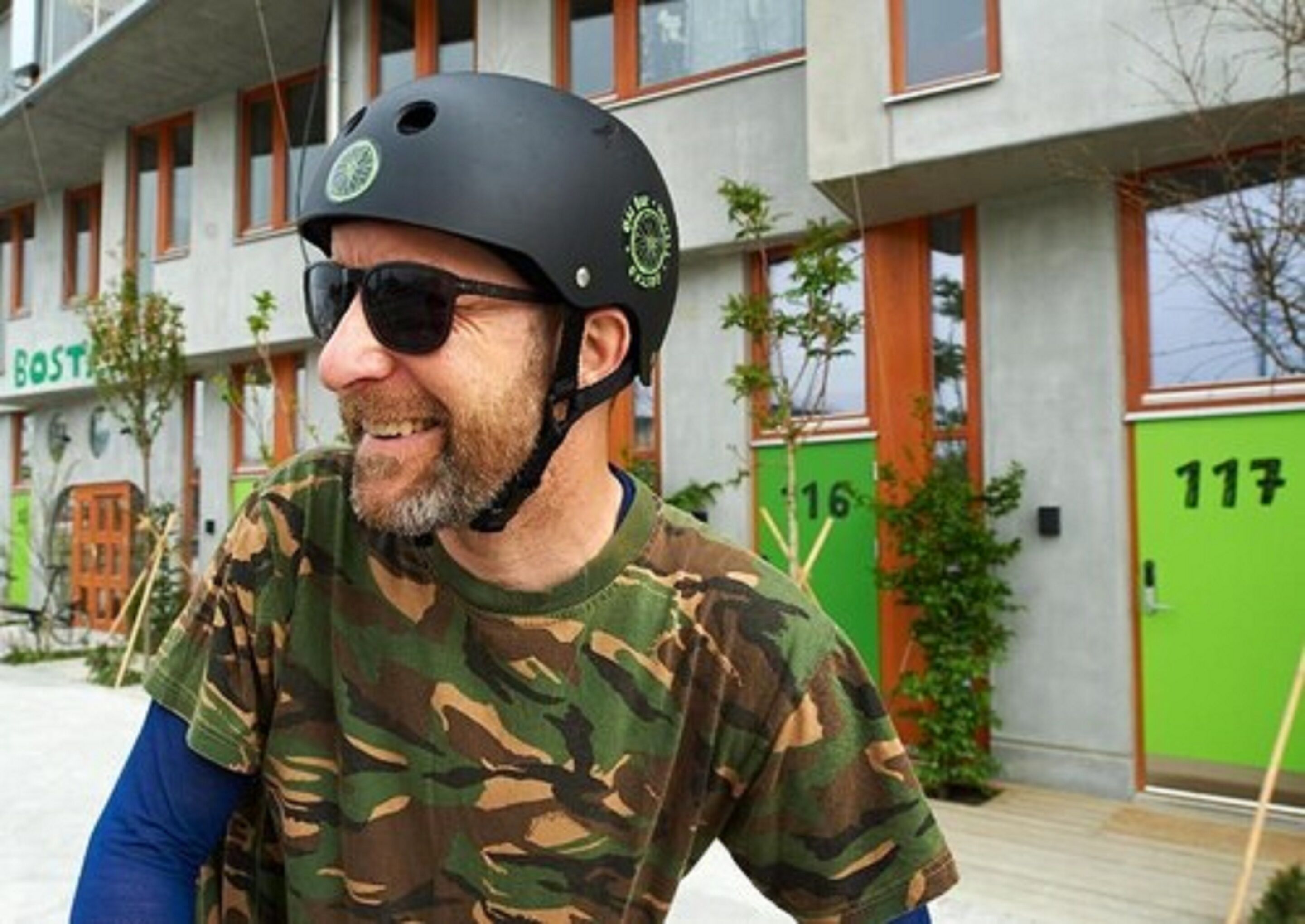 Una persona con casco y camisa de camuflaje de pie frente a un edificio con una puerta verde con el número 117.