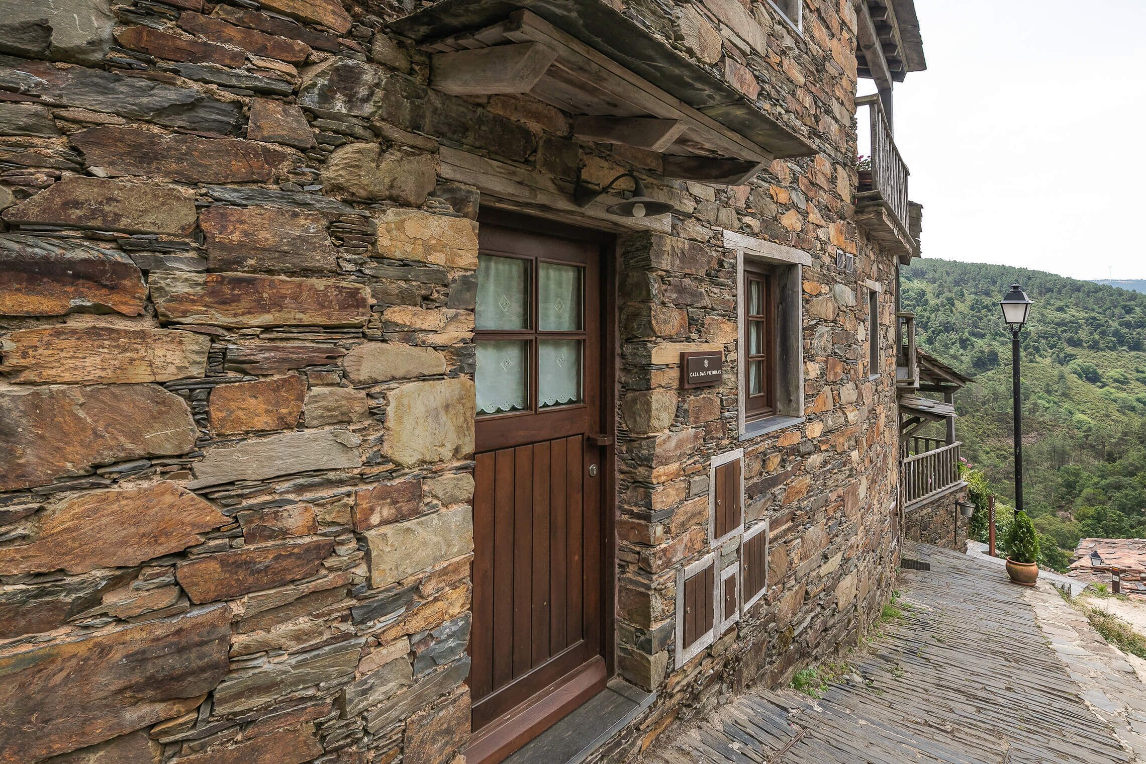 Un edificio de piedra con puerta y ventanas de madera, un balcón con planta en maceta y vistas a una colina boscosa.