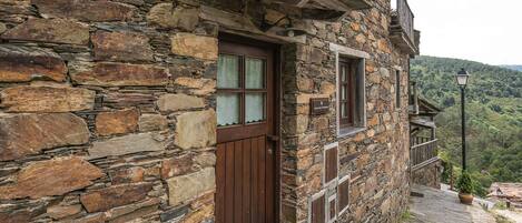 Un edificio de piedra con puerta y ventanas de madera, un balcón con planta en maceta y vistas a una colina boscosa.