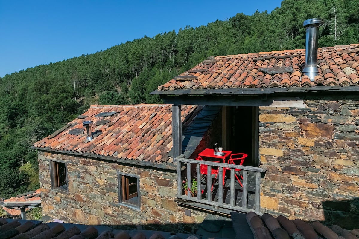 A stone building with a tiled roof, a balcony with red chairs, and a chimney.