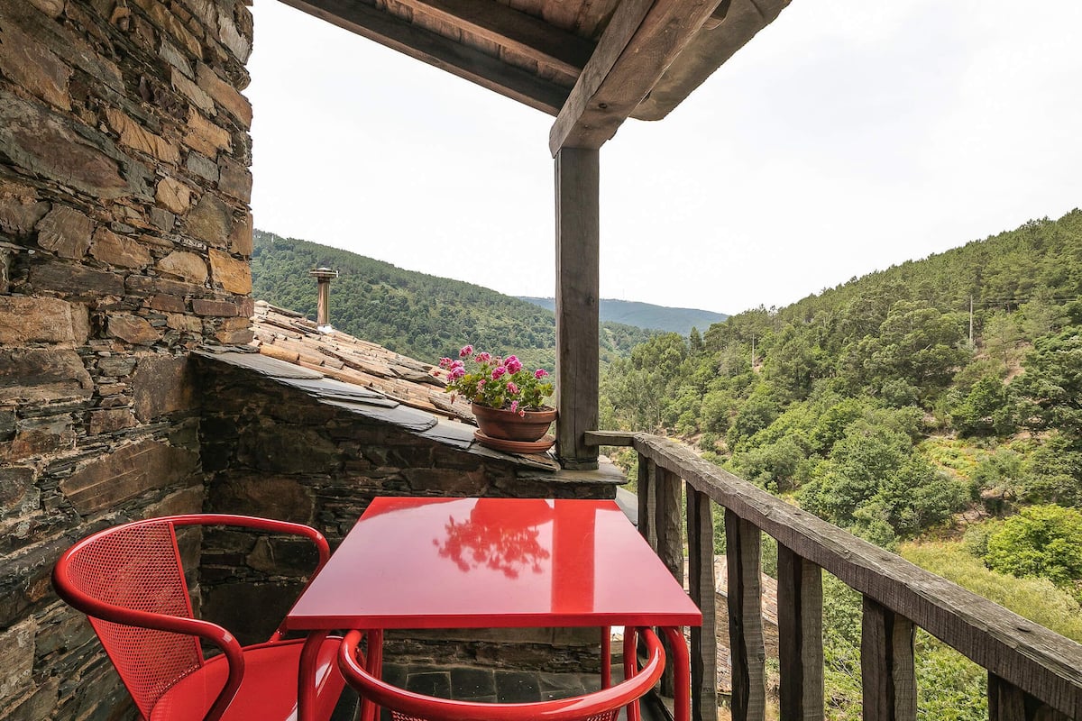 A stone building with a red table and chairs, a wooden railing, and a view of a forested hillside.