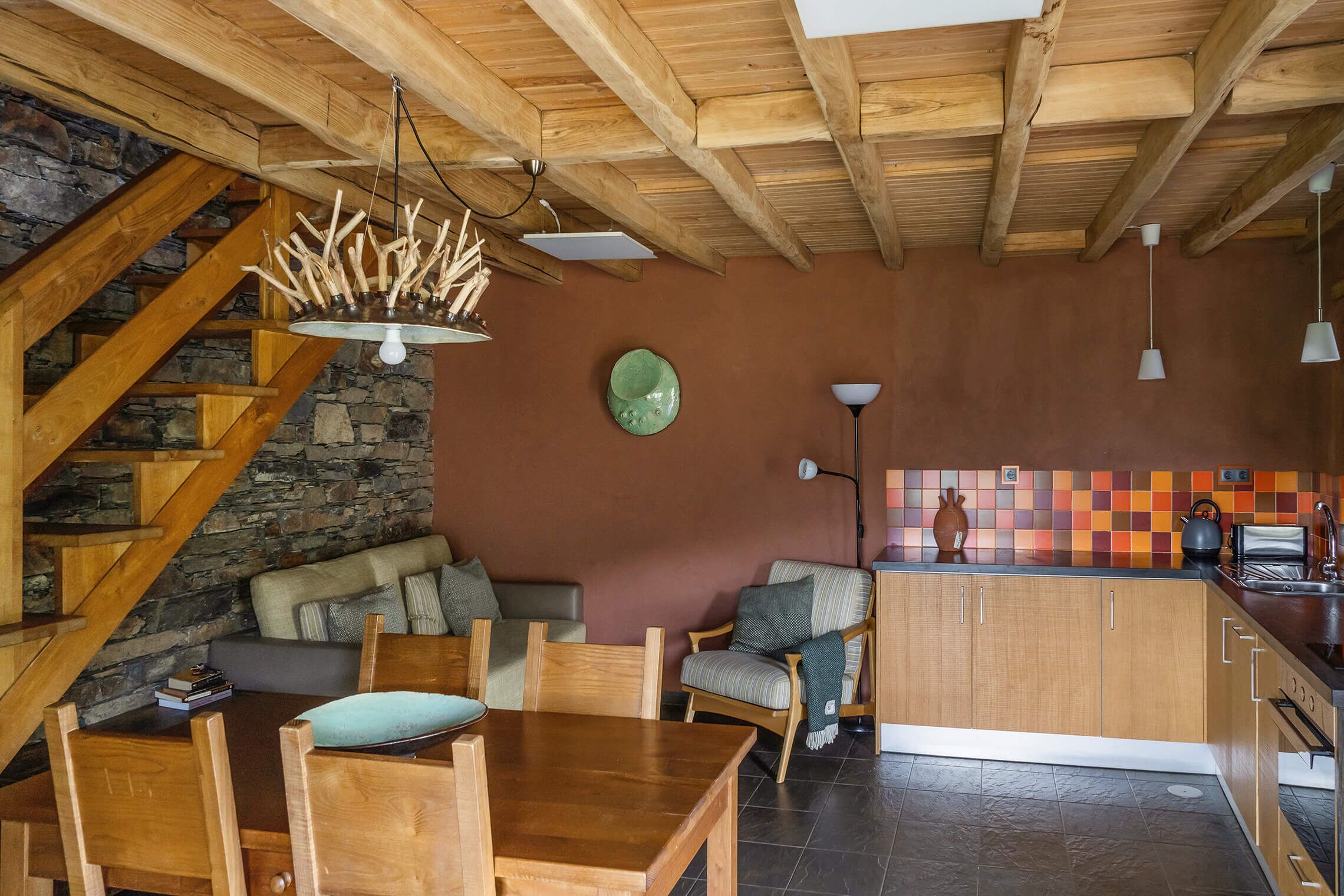 Uma mesa de jantar de madeira com cadeiras, uma área de cozinha com bancada e armários, e uma escadaria de pedra.