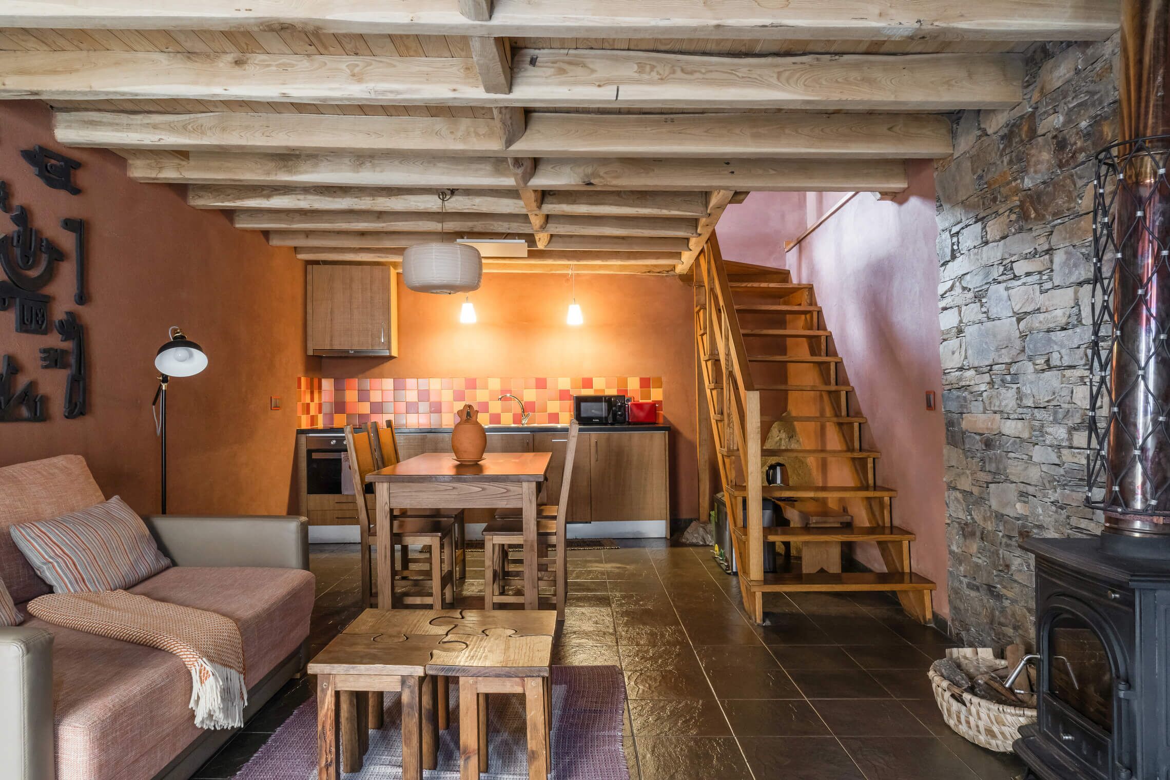 A cozy living area with a sofa, wooden table, and a fireplace. A kitchen with a stone wall and wooden staircase is visible in the background.