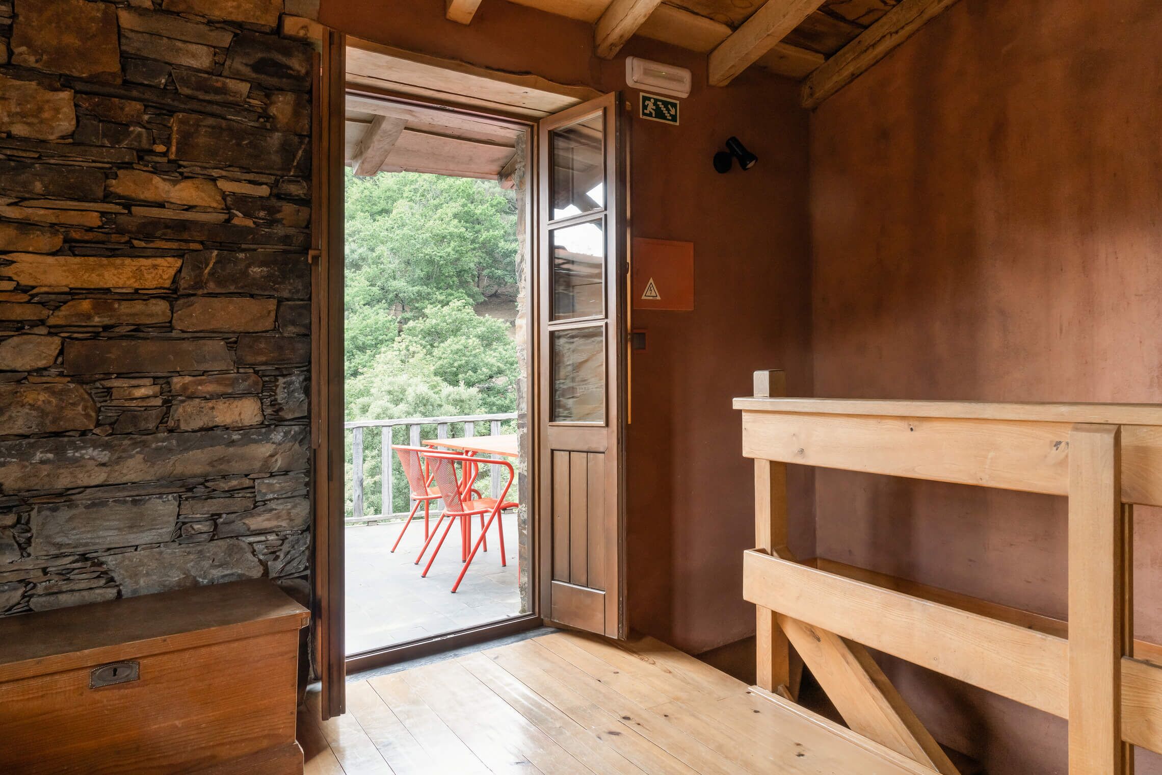 Interior de madera con una pared de piedra, un banco de madera, una puerta que da a un área exterior con sillas rojas y un estante de madera.