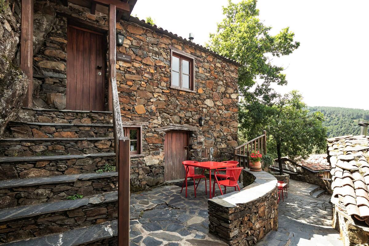 A stone building with a wooden door and window, outdoor seating area with red chairs, and a stone staircase.