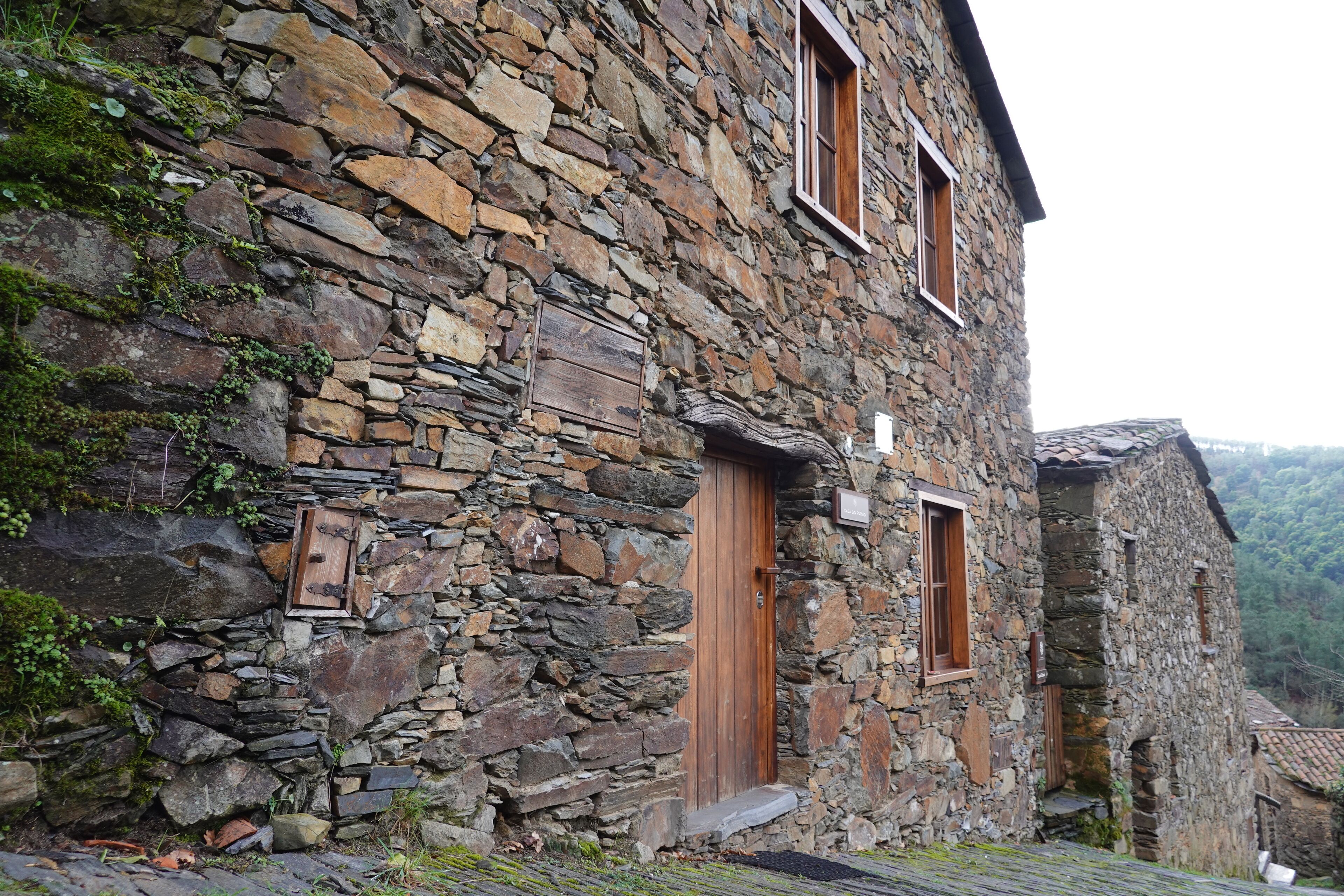 A stone building with wooden doors and windows, surrounded by greenery.