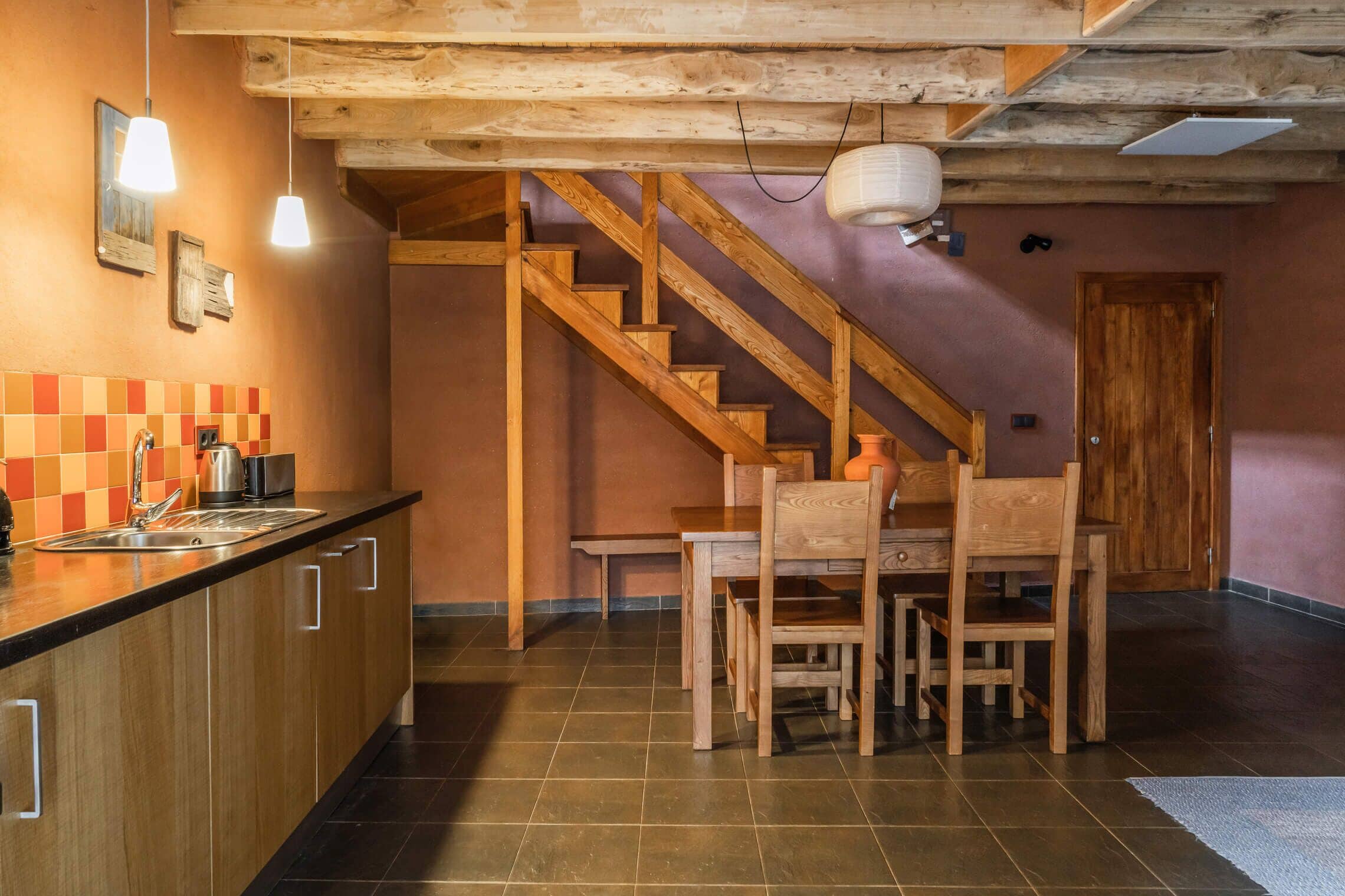 A kitchen with wooden cabinets, a black countertop, and a tiled backsplash. There is a wooden dining table with chairs and a staircase leading to an upper level.