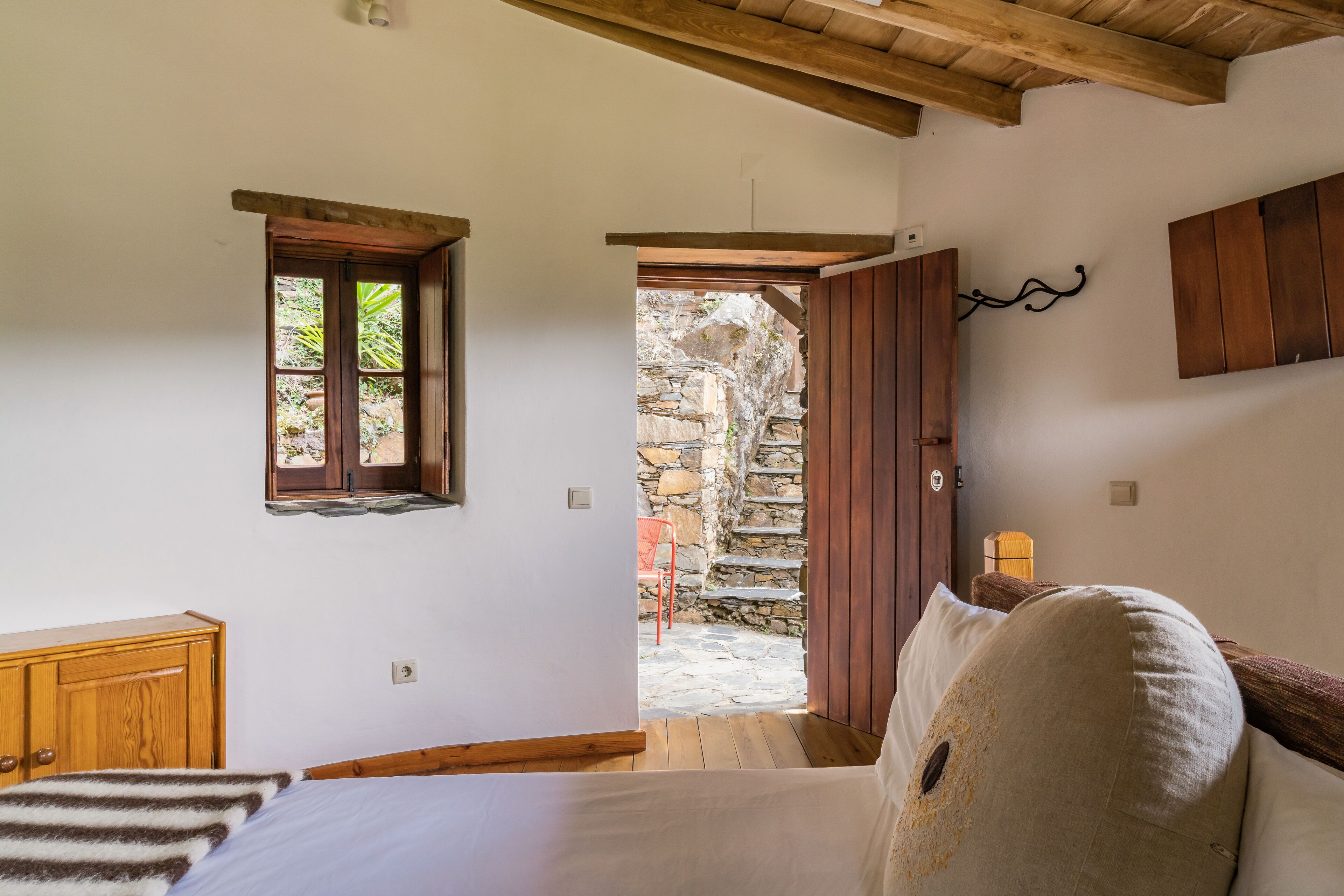 A bedroom with a bed, wooden headboard, a window, a small wooden cabinet, and a stone staircase leading outside.