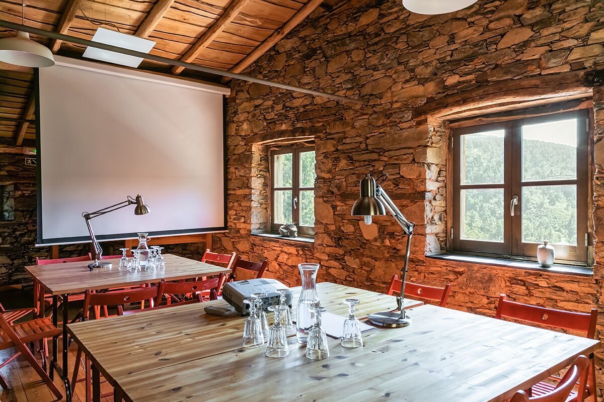 A rustic room with stone walls, wooden tables, and red chairs. There is a projector screen and a window with a view of trees.