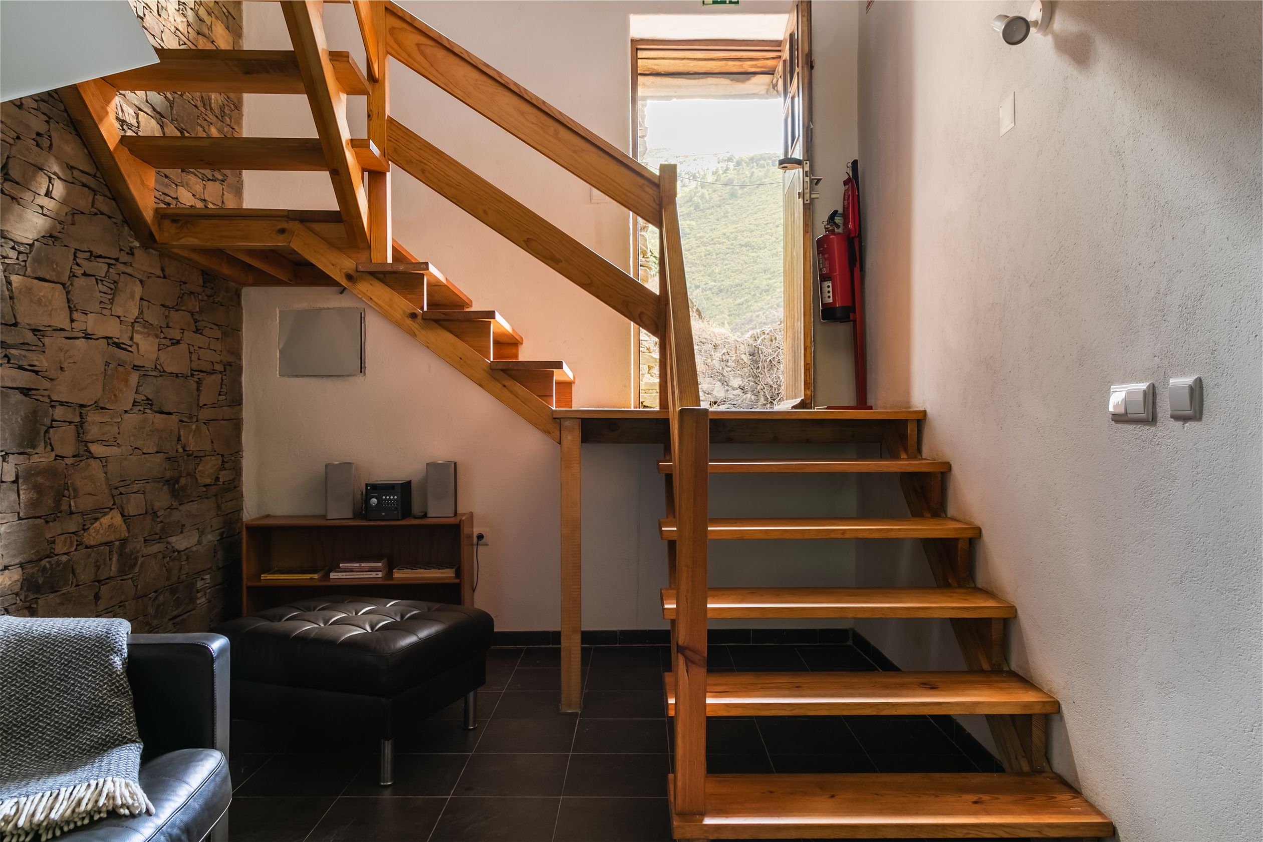 A staircase with wooden steps leading to a landing, a stone wall, a black leather sofa, and a small shelf with books and a speaker.