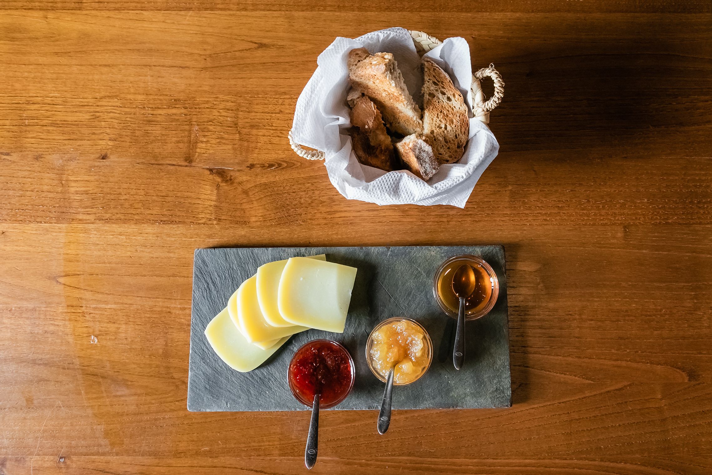 A wooden table with a slate serving board holding cheese and jams, and a basket of bread.