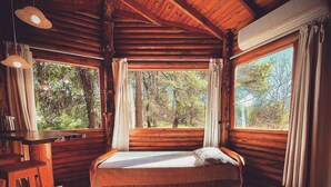 A log cabin room with a bed, wooden walls, and large windows offering a view of trees. - Posada del Viento (Tandil)