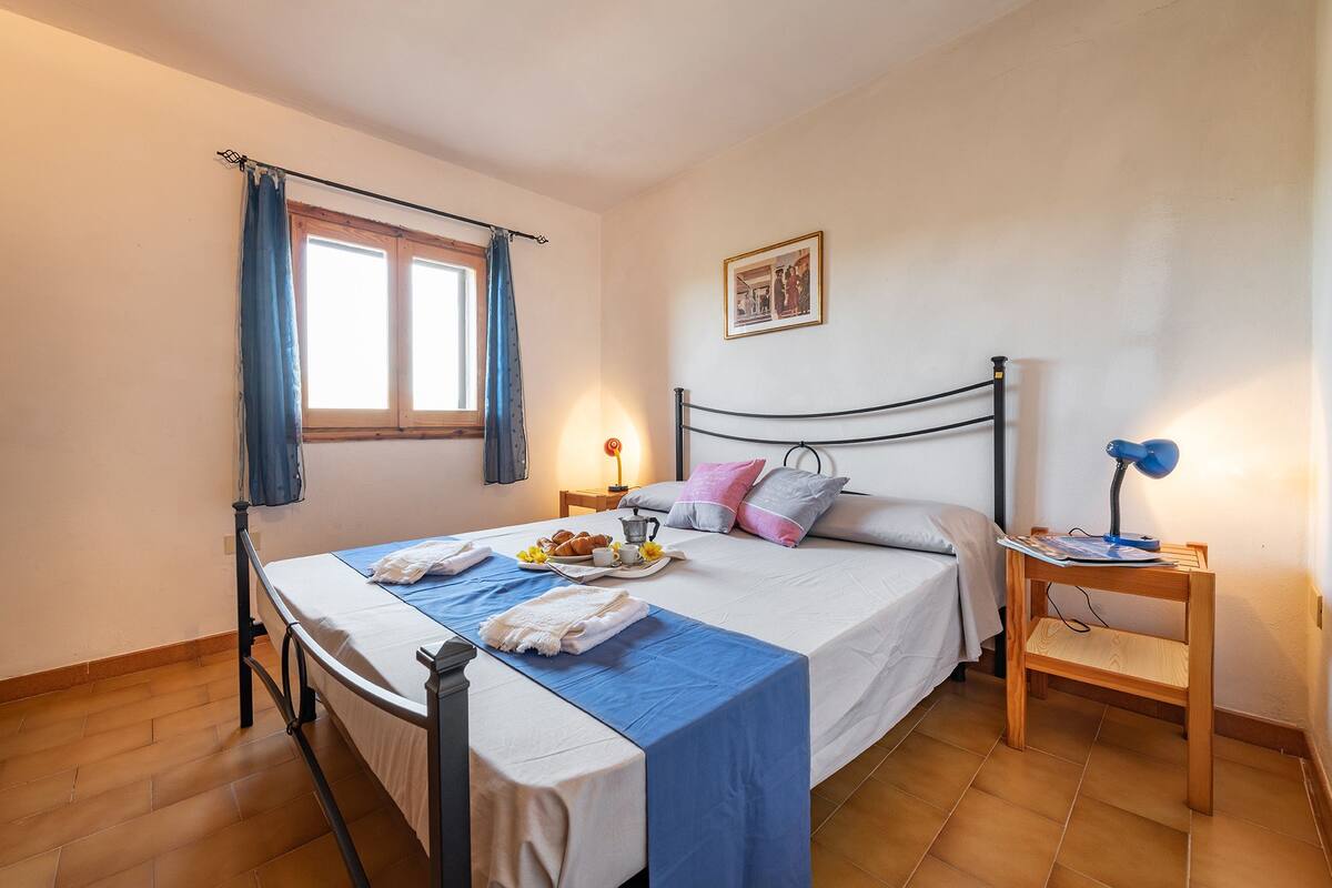 Un dormitorio ordenado con cama, mesita de noche, lámpara y ventana con cortinas.