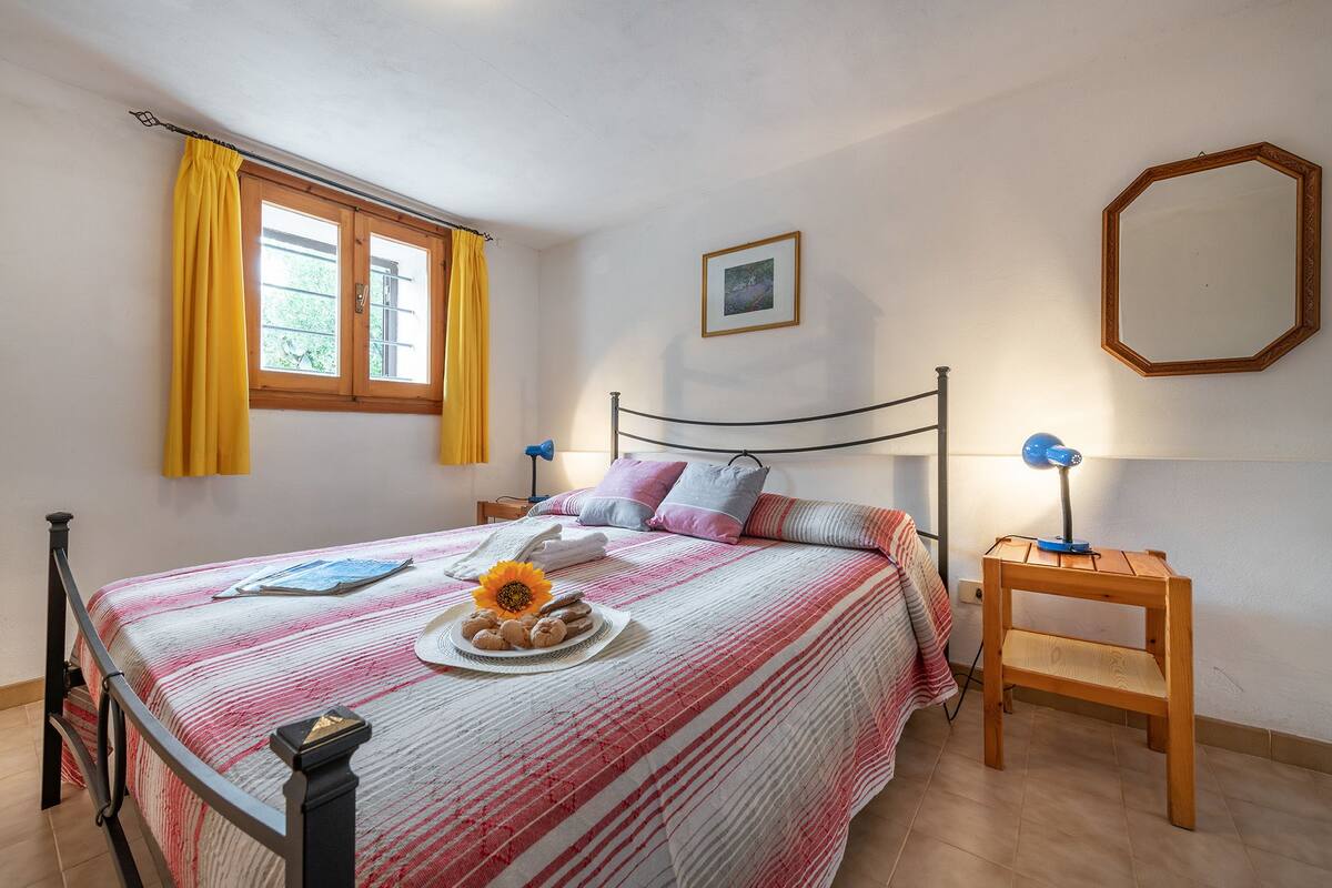 Un dormitorio con cama, mesita de noche, ventana con cortinas amarillas, un cuadro en la pared y un espejo.