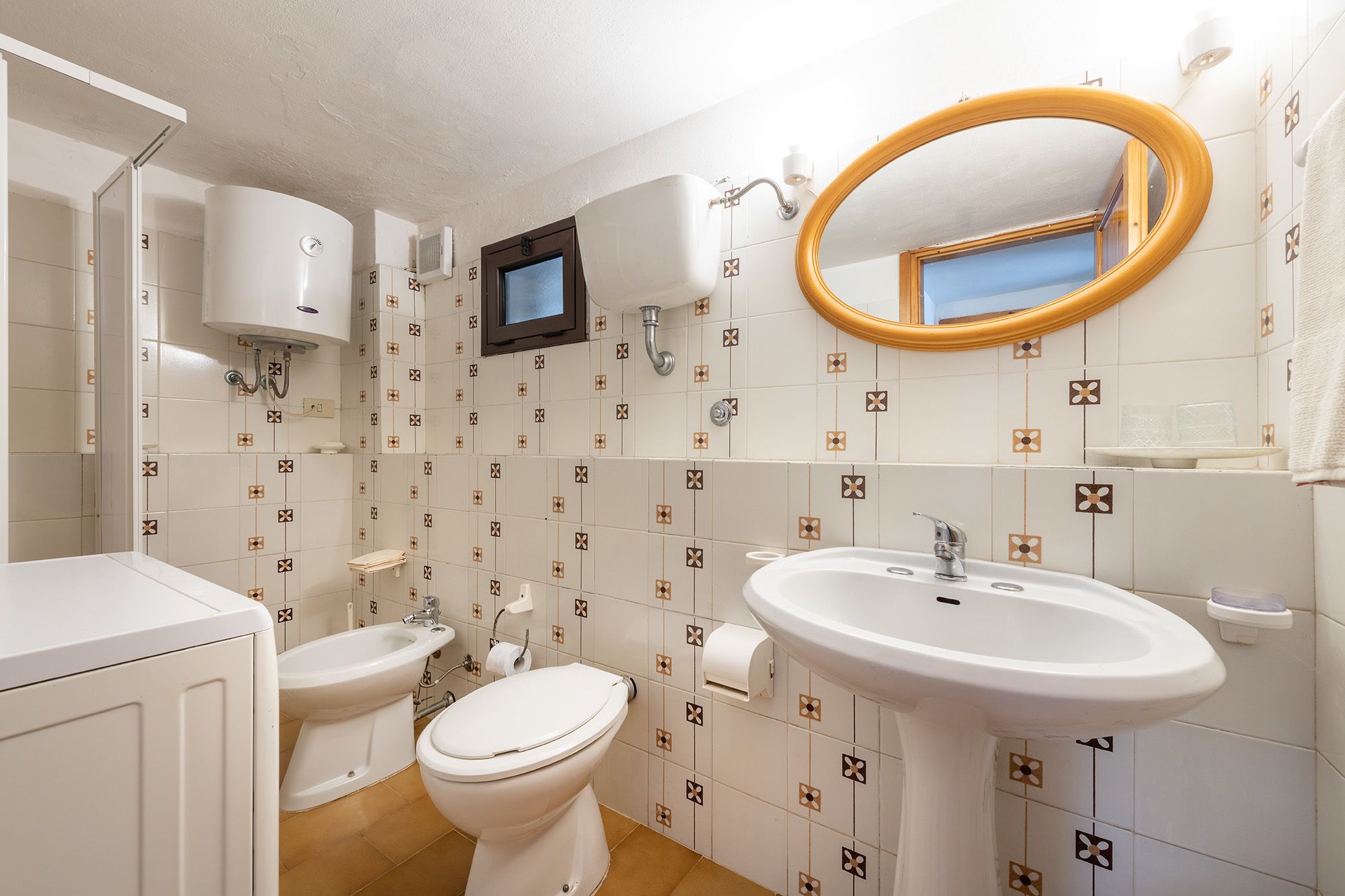 Un baño con inodoro, bidé, lavamanos y espejo con ventana arriba.