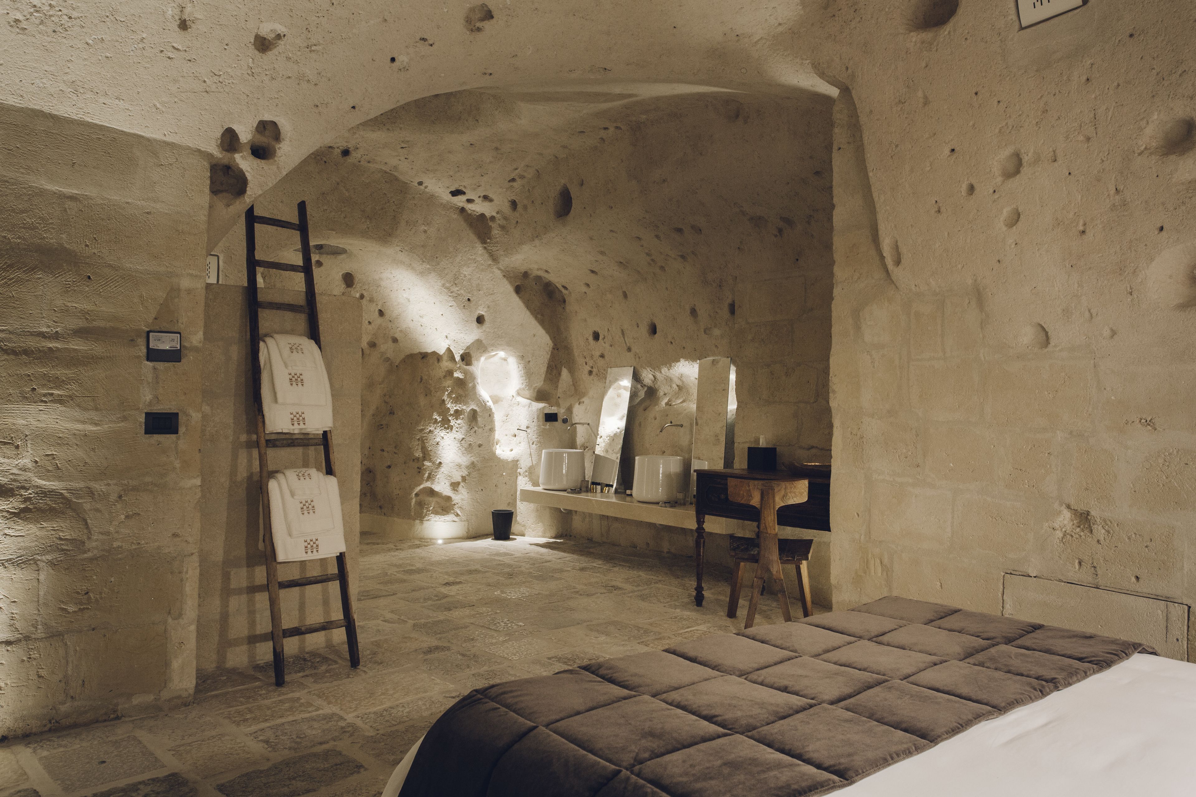 A rustic stone room with a bed, a wooden ladder, a desk, and a chair.