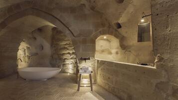 Ein Badezimmer mit Steinwänden, einer freistehenden Badewanne, einem kleinen Hocker und einem Spiegel an der Wand.