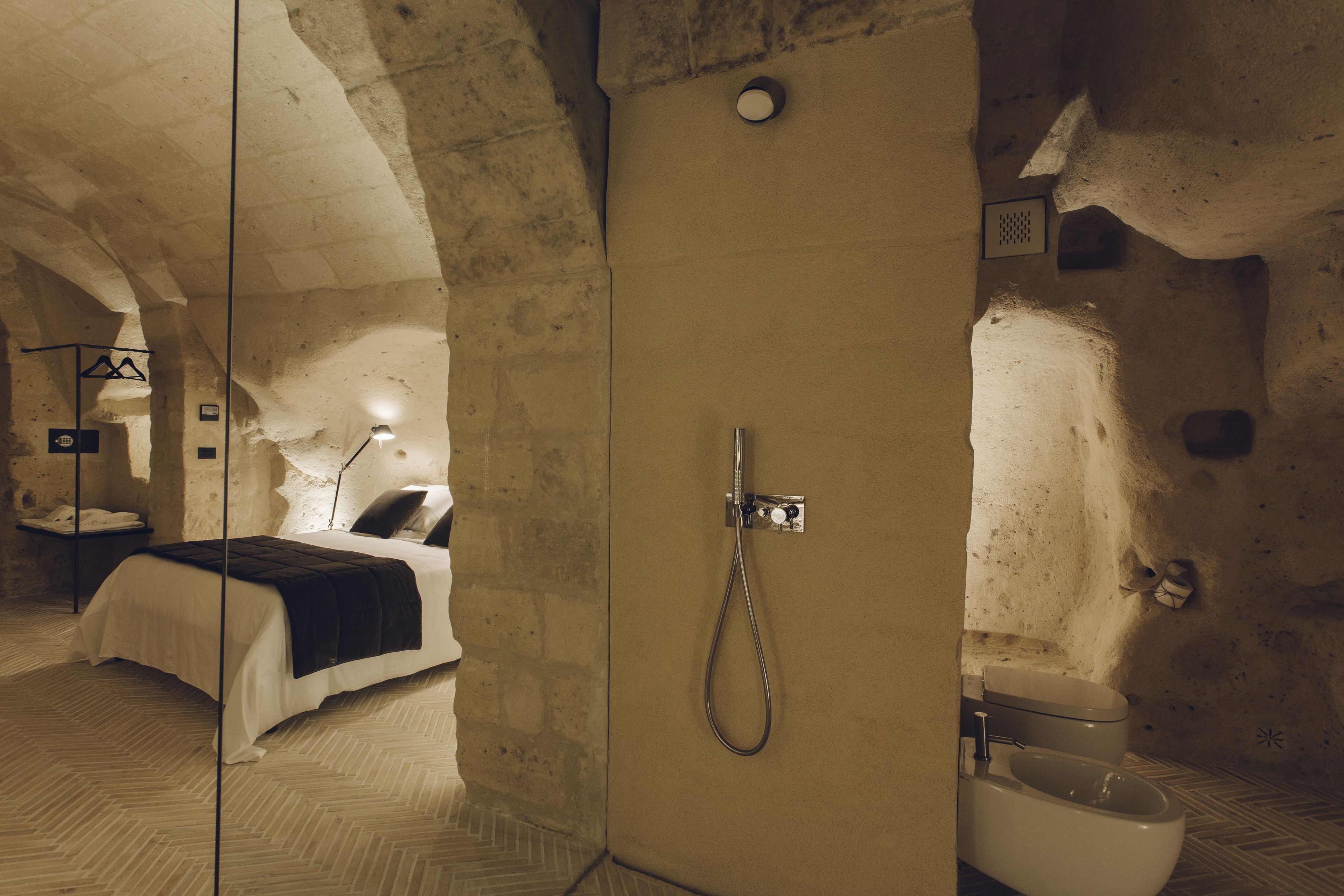 A stone-walled bathroom with a shower, toilet, and a bed with a white and black bedspread.