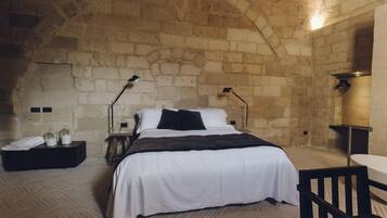 A bedroom with a stone wall, a bed with white bedding, two bedside lamps, a small table, and a chair.