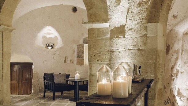 An interior with stone walls, a wooden bench, a table with candles, and a wooden cabinet. - Il Palazzotto (Matera)