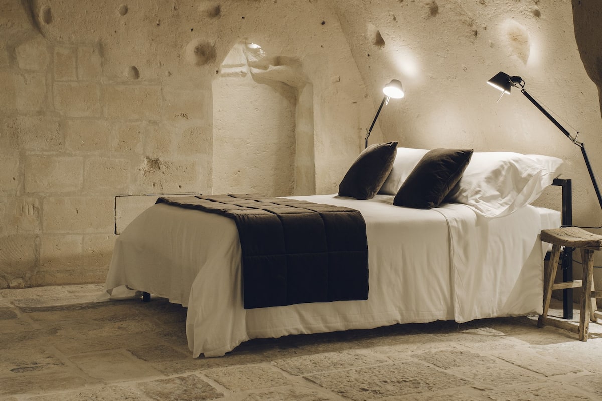 A bedroom with a stone wall, a bed with white bedding, two pillows, a bedside table, and a floor lamp.
