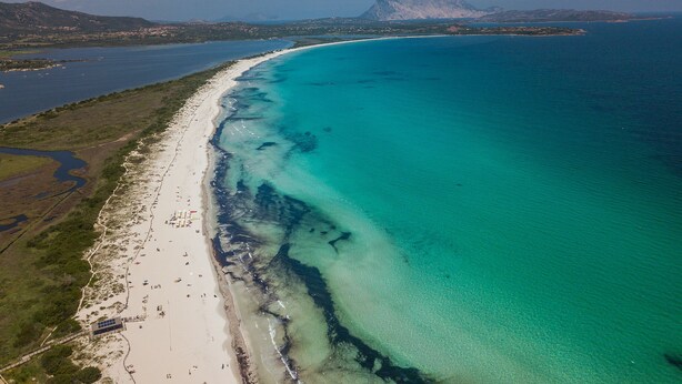 Vista aérea de una playa costera con aguas turquesas cristalinas, una orilla arenosa y una cadena montañosa distante.