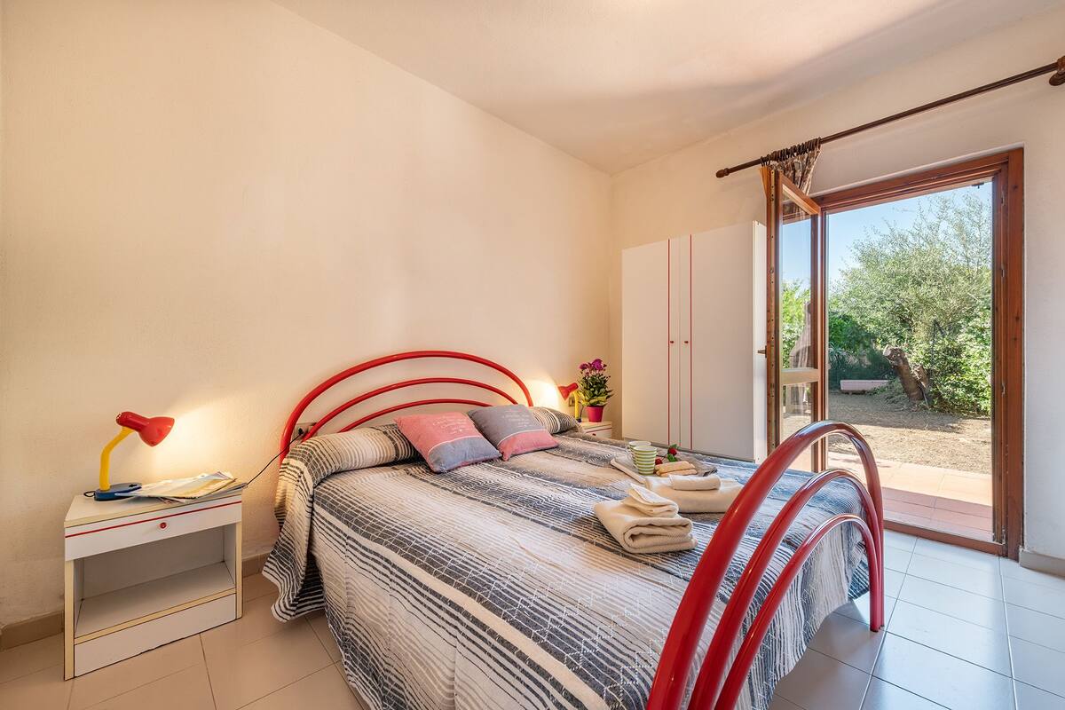 Ein Schlafzimmer mit Bett, Nachttisch, Lampe, Kleiderschrank und einer Schiebeglastür zum Garten.