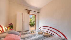 Un dormitorio con cama, mesita de noche con lámpara, planta en maceta y puerta que da a un área exterior.