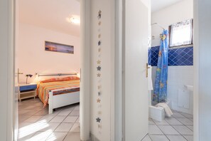 A bedroom with a bed, bedside table, and a nightstand. There is a bathroom with a shower, toilet, and sink.