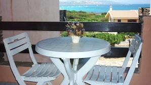 Un balcon avec une table ronde et deux chaises blanches, offrant une vue sur la côte, avec des bâtiments et un ciel dégagé.