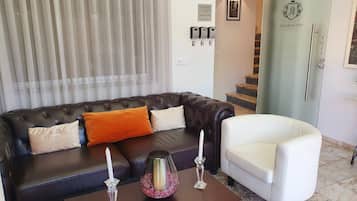 Uma sala de estar com um sofá de couro marrom escuro, uma poltrona branca e uma mesa de centro com velas e um vaso decorativo.