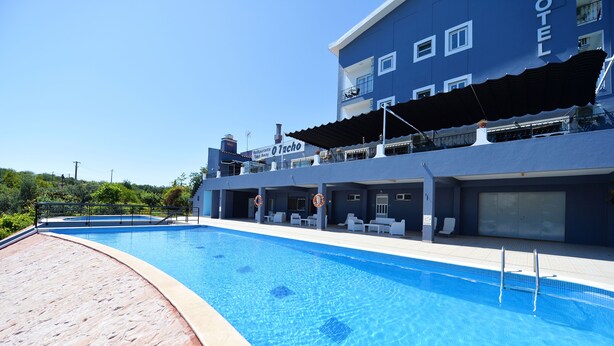 A swimming pool with a blue-tiled deck, a hotel building with a sign reading "O Tacho Restaurant Bike Andy," and a clear sky. - Hotel Don Rodrigues (Tavira)