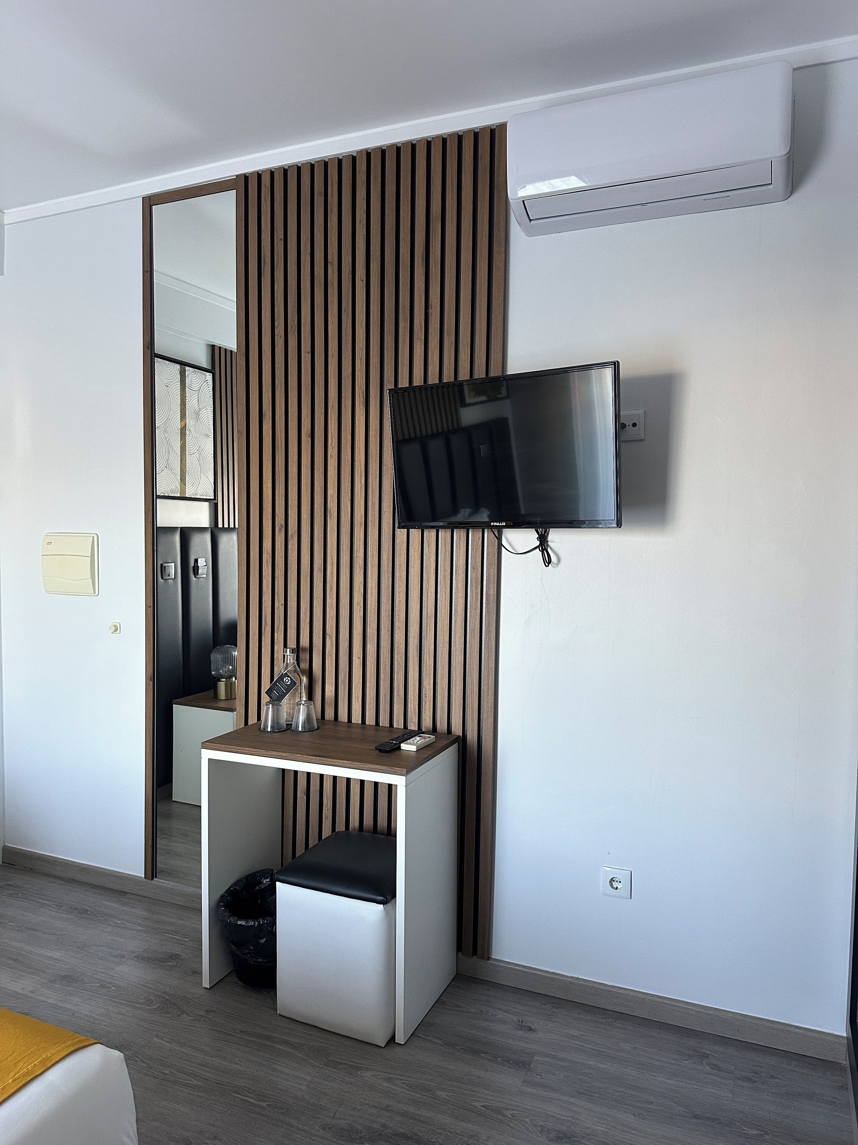 A modern hotel room with a wooden panel wall, a mounted flat-screen TV, a small bedside table with a lamp, and a trash bin.