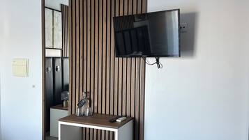 A modern hotel room with a wooden panel wall, a mounted flat-screen TV, a small bedside table with a lamp, and a trash bin.