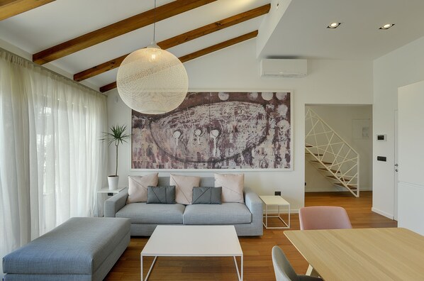 A modern living room with a grey sofa, a white coffee table, and a wooden dining table. There is a large abstract painting on the wall and a staircase in the background.