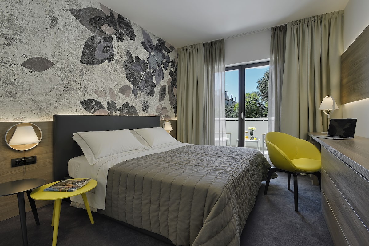 A hotel room with a large bed, a desk with a laptop, a yellow chair, and a view of the outdoors.