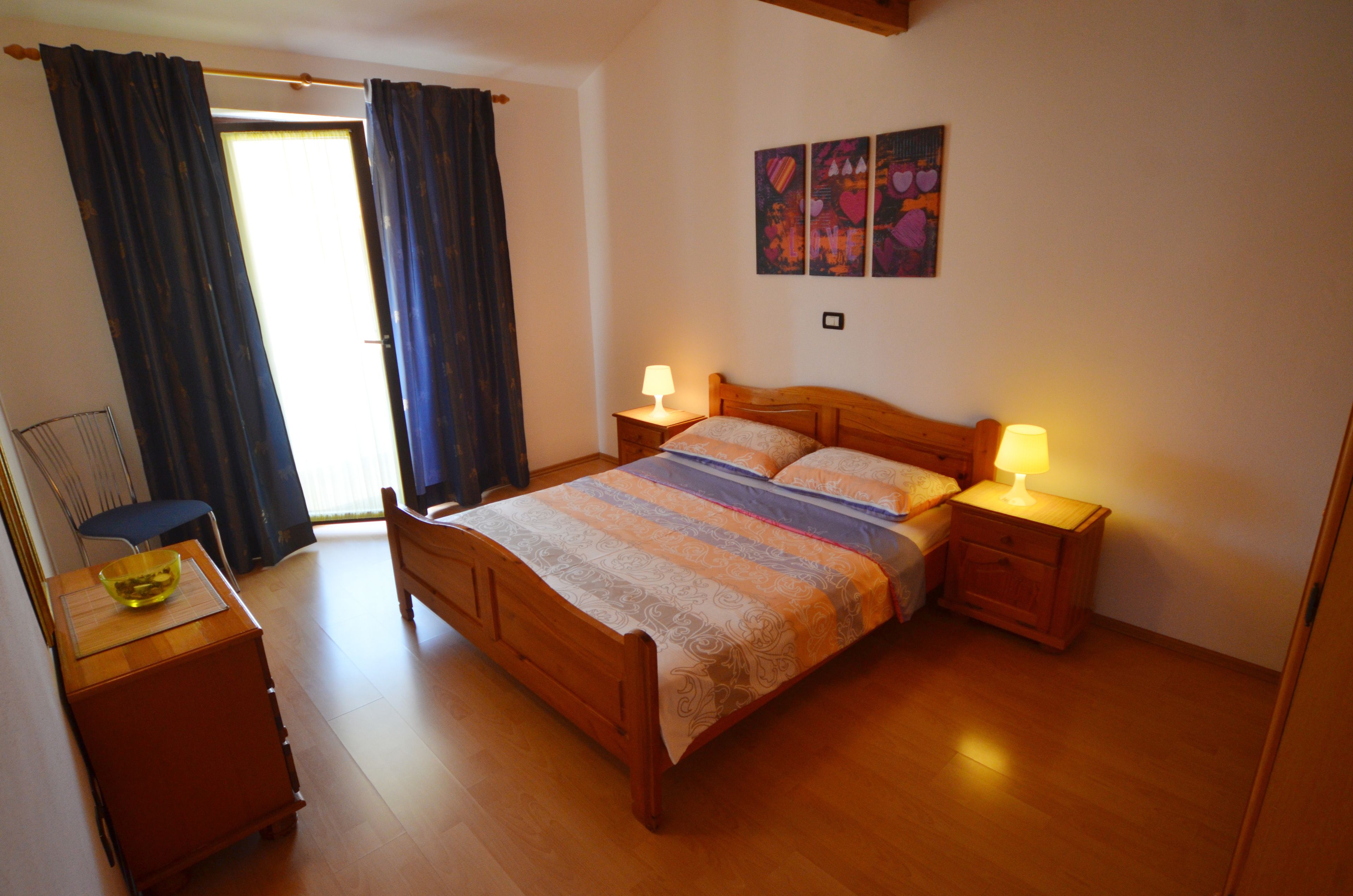 A bedroom with a wooden bed, two bedside tables with lamps, a chair, and a window with curtains.